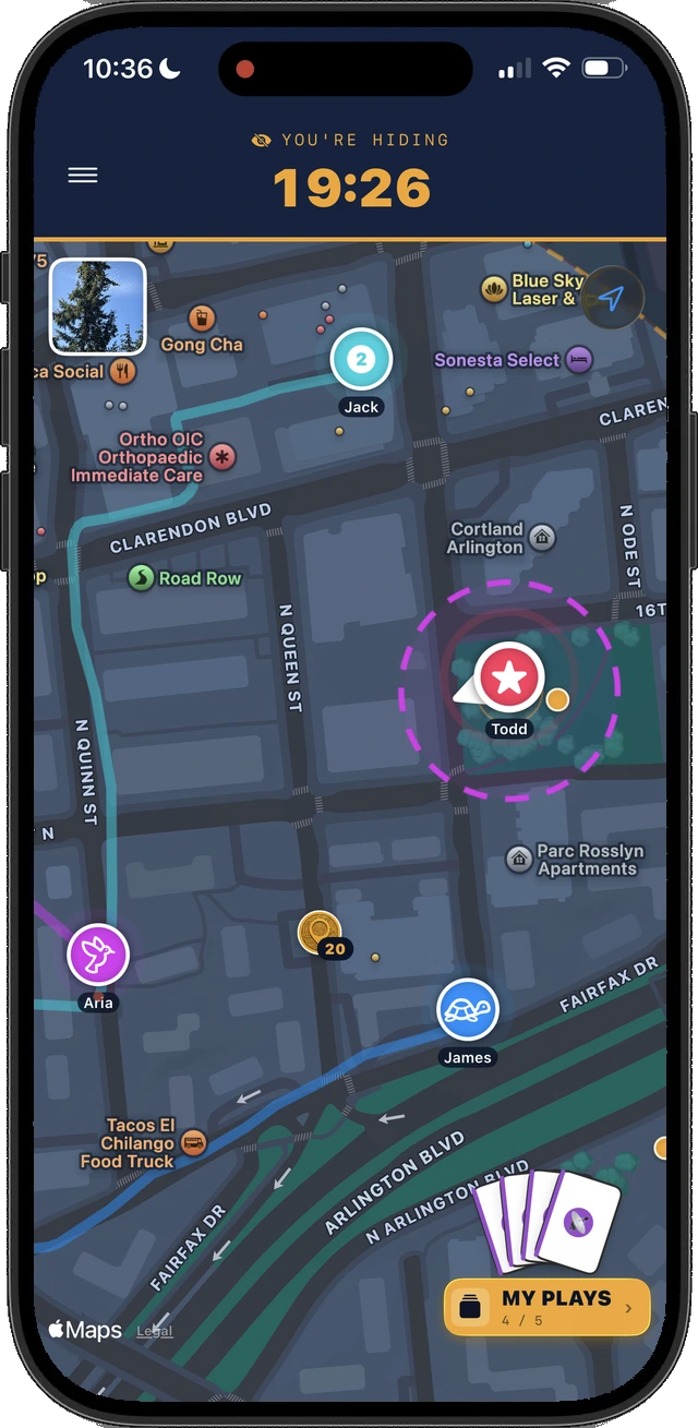 Phone screen showing the in-round map from the Hider's own device. A banner reads YOU'RE HIDING above a 19 minute 26 second clock, Seeker pins move along the streets, coin pickups sit on the map, and the Hider's own position is marked inside a dashed circle.